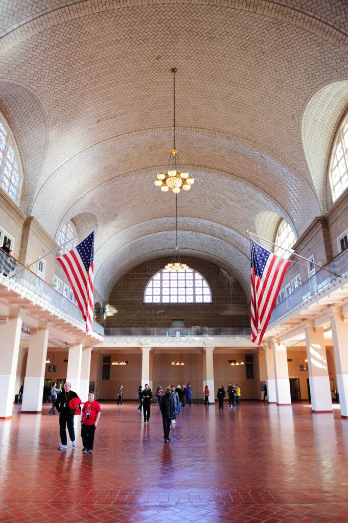 Grand indoor hall with arched, tiled ceiling, chandelier, and American flags along the balcony; visitors on a red-tiled floor.